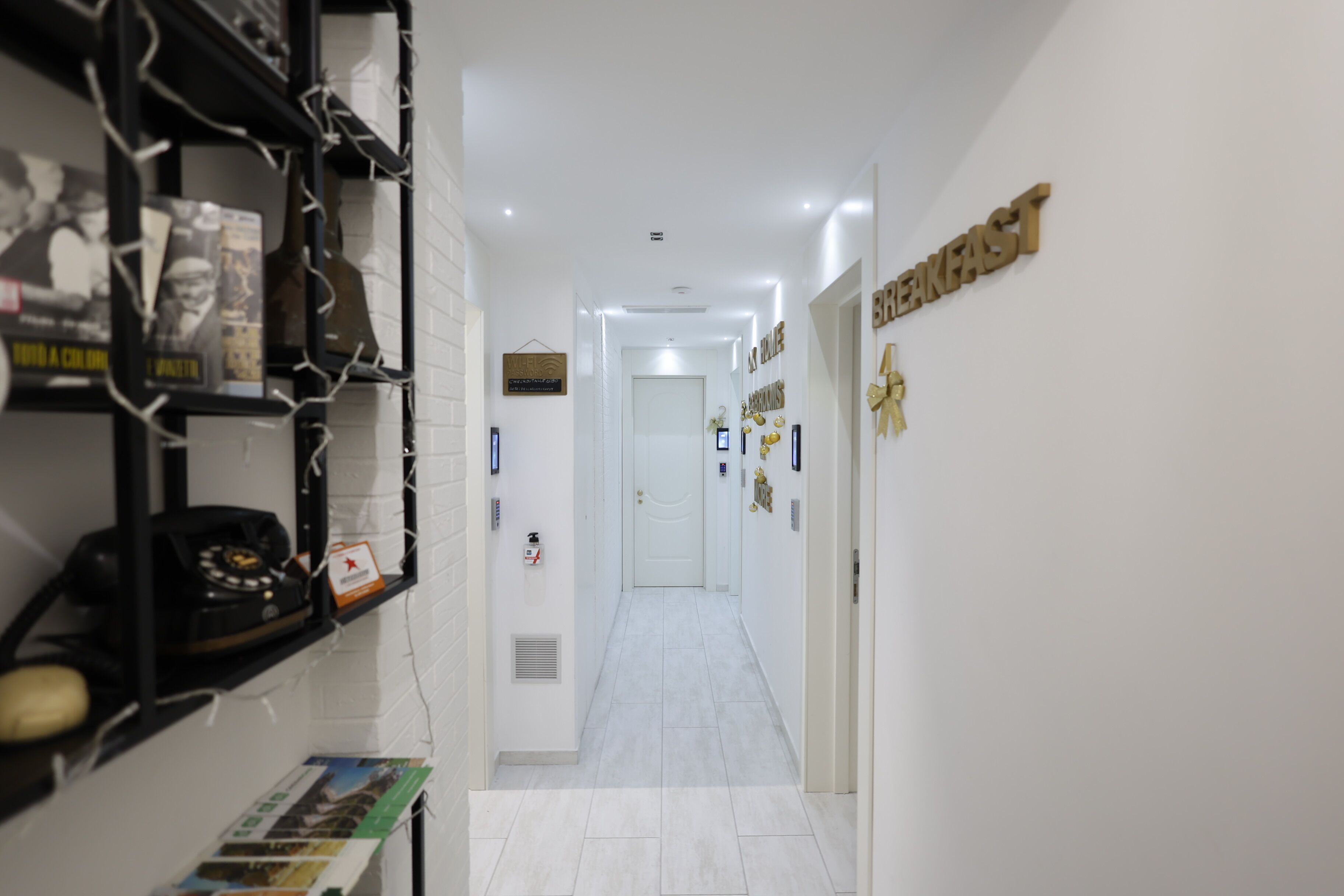 A hallway with a white tiled floor, a door at the end, and a shelf with various items including a telephone and a box.