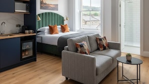 A modern hotel room with a bed, a sofa, a small table, a kitchenette, and a large window offering a view of the outside. - The Civic Hall - Wayth Hotels (Hebden Bridge)