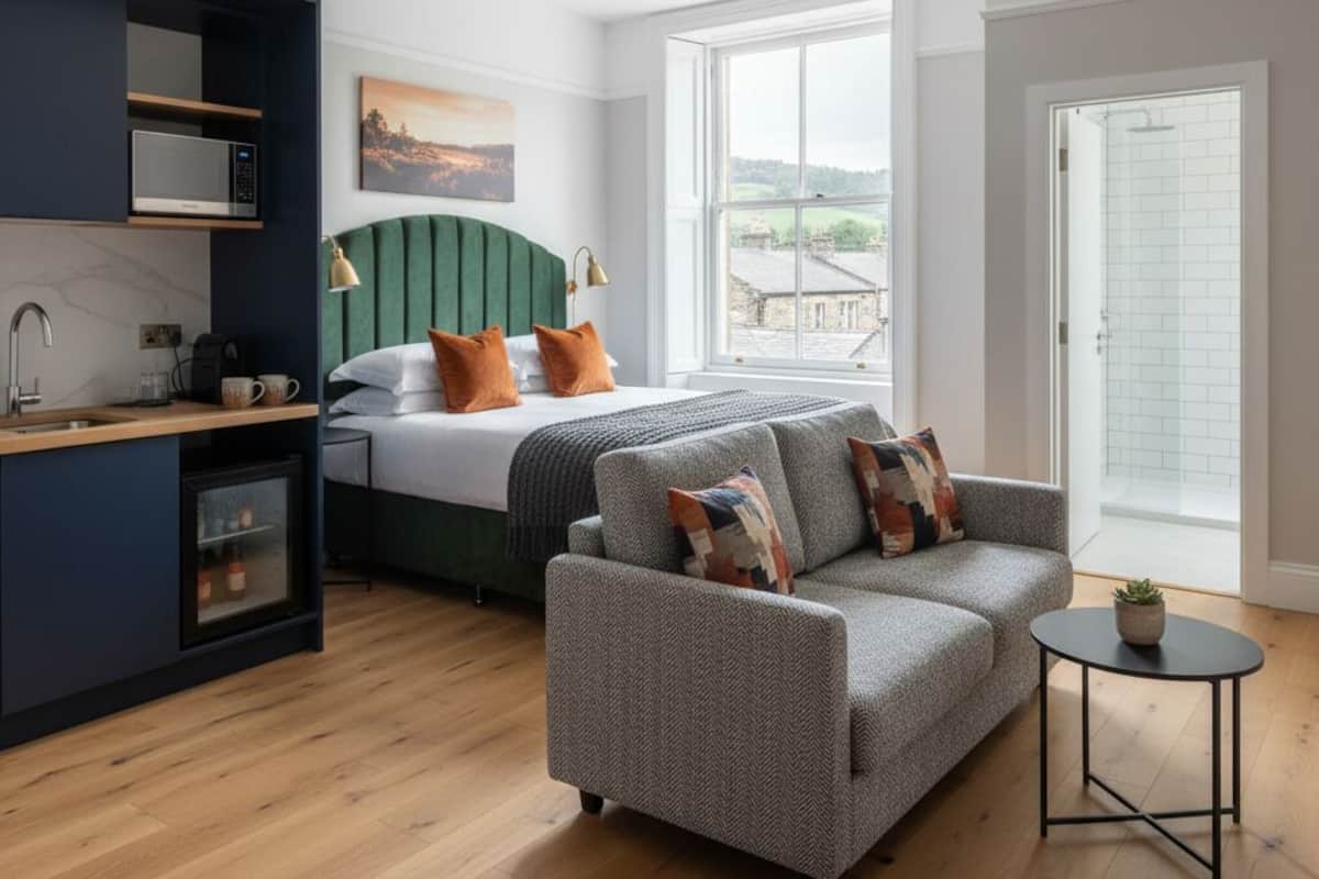 Una habitación de hotel moderna con cama, sofá, una mesita, kitchenette y un gran ventanal con vistas al exterior.