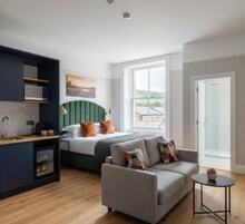 Una habitación de hotel moderna con cama, sofá, una mesita, kitchenette y un gran ventanal con vistas al exterior.