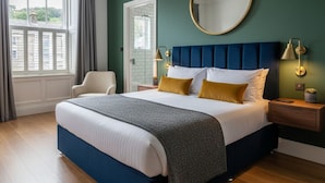 A bedroom with a large bed, a round mirror, a nightstand with a lamp, and a view of a green landscape through a window. - The Civic Hall - Wayth Hotels (Hebden Bridge)