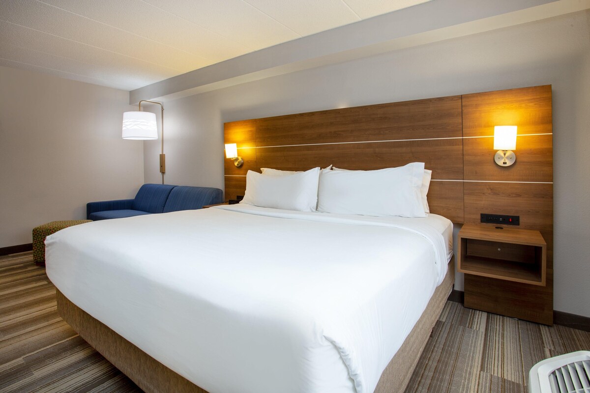 Habitación de hotel con una cama grande, una mesita de noche con lámpara, una silla azul y un pequeño reposapiés.