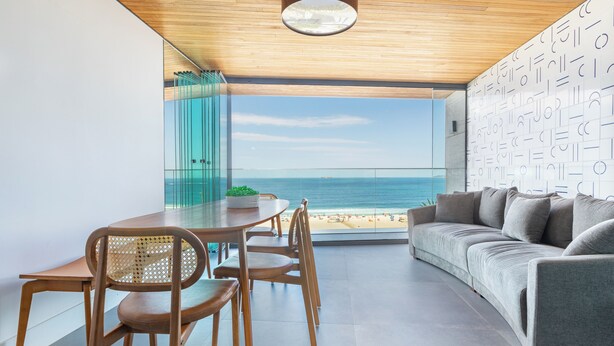 A modern living room with a large window overlooking a beach, a wooden dining table with chairs, and a grey sofa. - Atlantica Prime Copacabana Ocean View (Rio de Janeiro)
