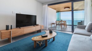 A modern living room with a flat-screen TV, wooden furniture, a blue carpet, and a view of the sea through large glass doors. - Atlantica Prime Copacabana Ocean View (Rio de Janeiro)