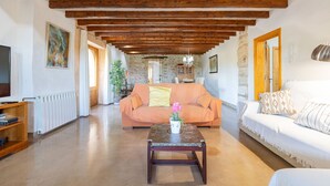 A spacious living room with exposed wooden beams, a sofa, a coffee table with a vase of flowers, and a television on a wooden stand.