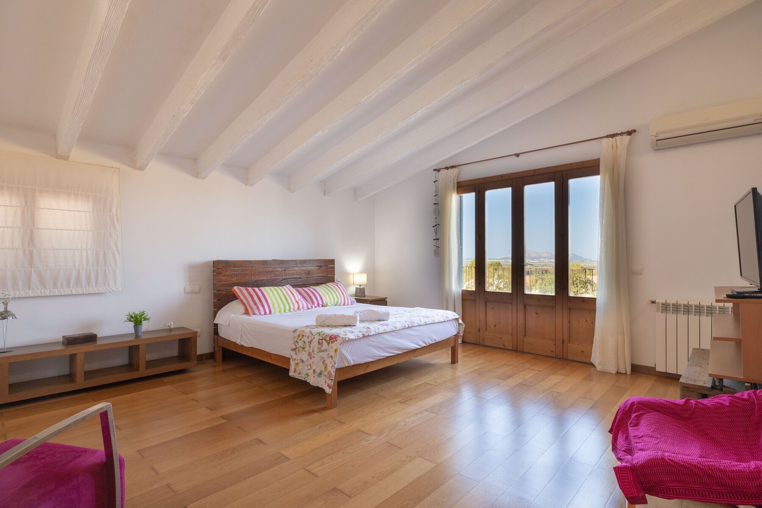 A bedroom with a wooden bed, a bench, a chair, and a view through a large window.