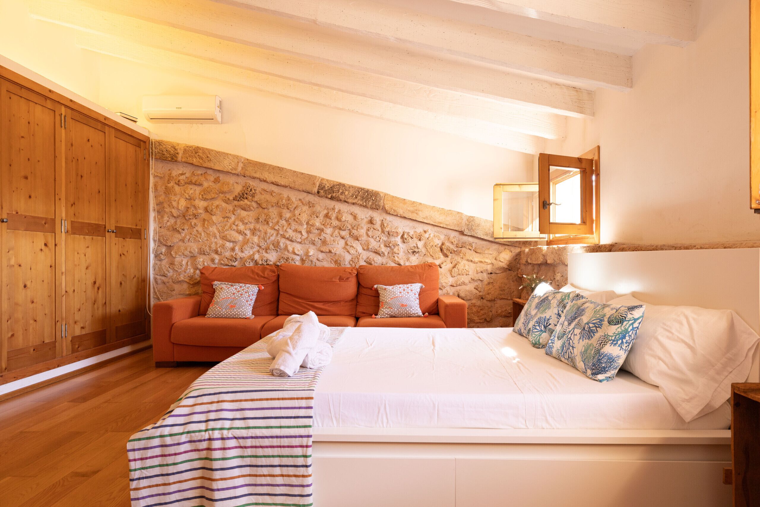 Ein Schlafzimmer mit einem Holzschrank, einem orangefarbenen Sofa, einem Bett mit weißer Bettwäsche und einer Steinwand.
