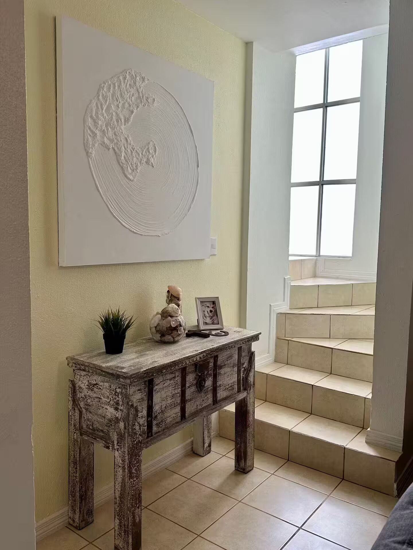 A room with a rustic wooden console table, a potted plant, and a framed picture. A large abstract artwork is mounted on the wall above the table.