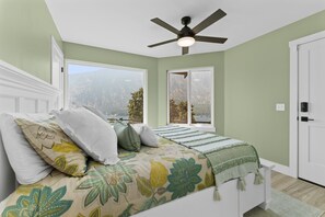 A bedroom with a bed, a ceiling fan, and two windows with mountain views.