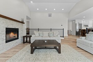 A spacious living room with a fireplace, a sofa, a coffee table, and a kitchen area in the background.