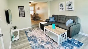 Un salon avec un canapé gris, une table basse et un tapis bleu. Au fond, on aperçoit un coin repas avec une table et des chaises.