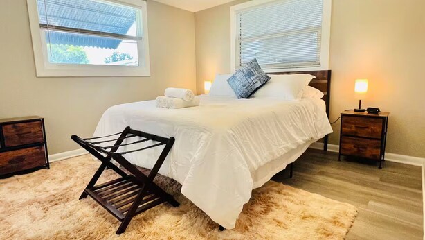 Un lit bien fait, avec du linge de lit blanc, un tapis beige moelleux et une chaise en bois. Il y a deux tables de chevet avec des lampes, une fenêtre avec des stores et un climatiseur fixé au mur.
