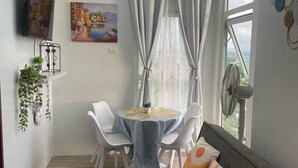 Un salon moderne avec un canapé gris, une table à manger avec des chaises, un téléviseur fixé au mur et une grande fenêtre avec des rideaux.