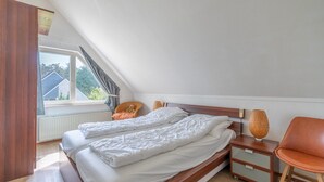 A bedroom with a sloped ceiling, a bed with white bedding, a nightstand with a lamp, a chair, and a window with a view of houses. - Charming Villa Near the sea and Forest (De Koog)