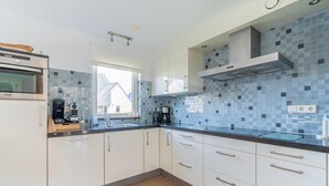 A modern kitchen with white cabinets, a built-in oven, a coffee maker, and a window providing natural light. - Charming Villa Near the sea and Forest (De Koog)