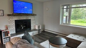 Una sala de estar con chimenea, televisor montado encima, un ventanal con vistas a follaje y un rincón de descanso acogedor con sofá y una mesa redonda.