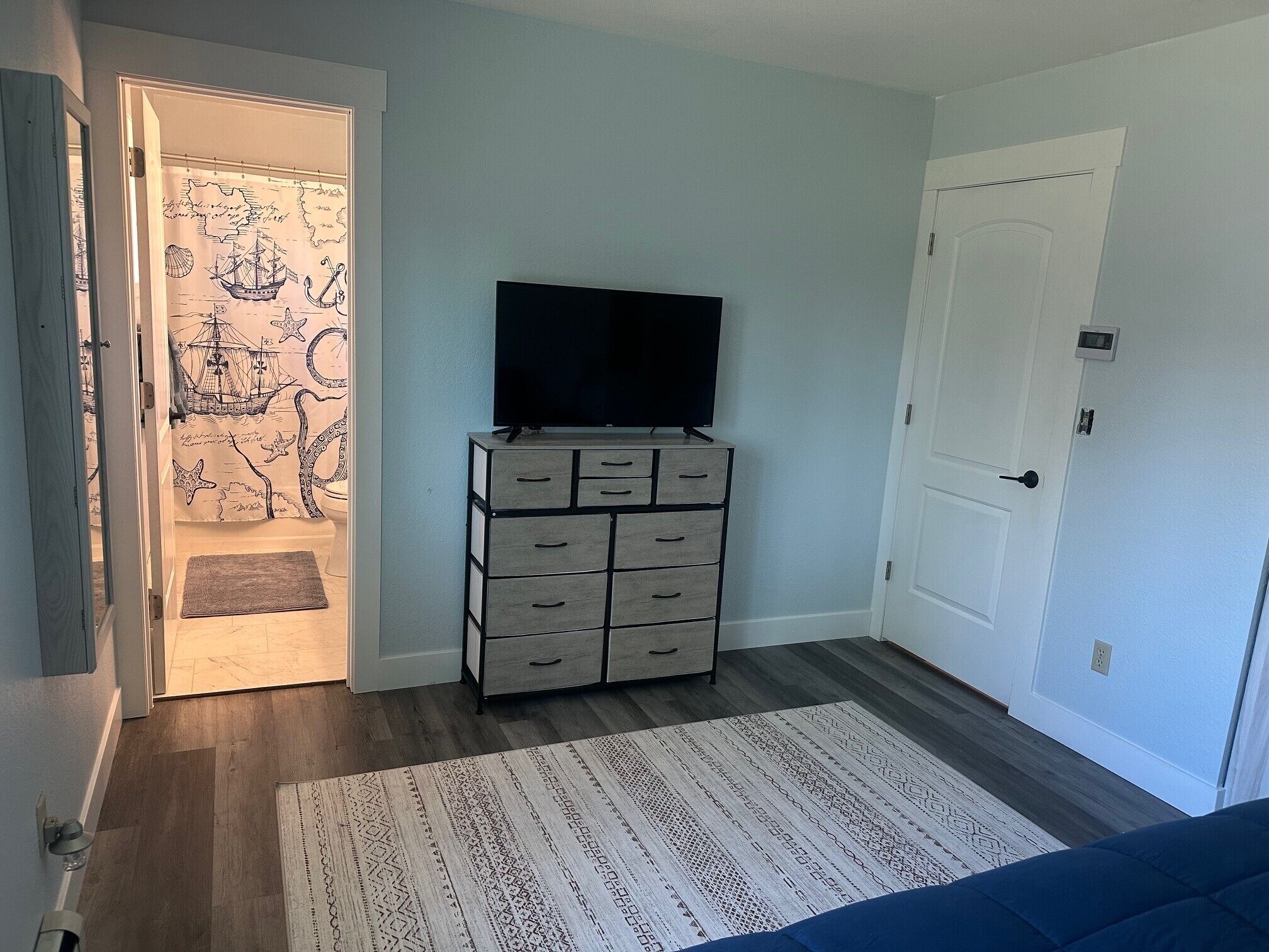 Ein Zimmer mit einem großen Flachbildfernseher auf einem Standfuß, einem gemusterten Teppich und einer Tür mit einem maritim designten Vorhang.