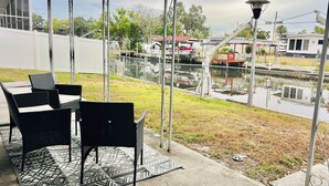 Eine Terrasse im Freien mit Tisch und Stühlen, einem gemusterten Teppich und Blick auf einen eingezäunten Bereich mit einem Boot und Gebäuden im Hintergrund.