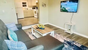 Ein Wohnzimmer mit grauem Sofa, einem Couchtisch mit Blumen, einem Fernseher an der Wand und einer Küche im Hintergrund.