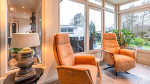 A modern living room with two orange leather armchairs, a side table with a lamp, and large windows offering a view of the outdoors.