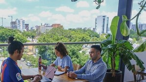 Drie personen zitten aan een houten tafel op een balkon met uitzicht over de stad.
