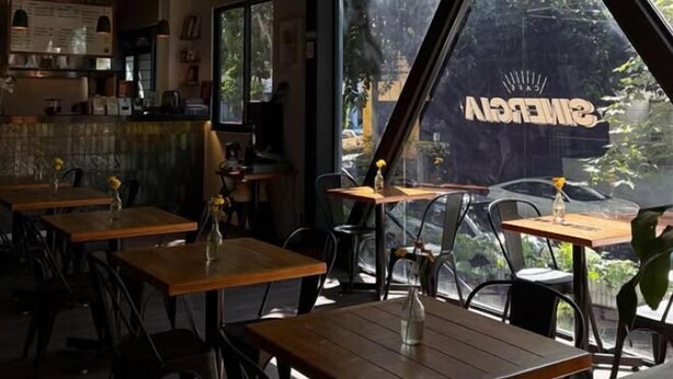 Een restaurantinterieur met houten tafels en stoelen, hangende pendantlampen en een groot raam met uitzicht op groen.