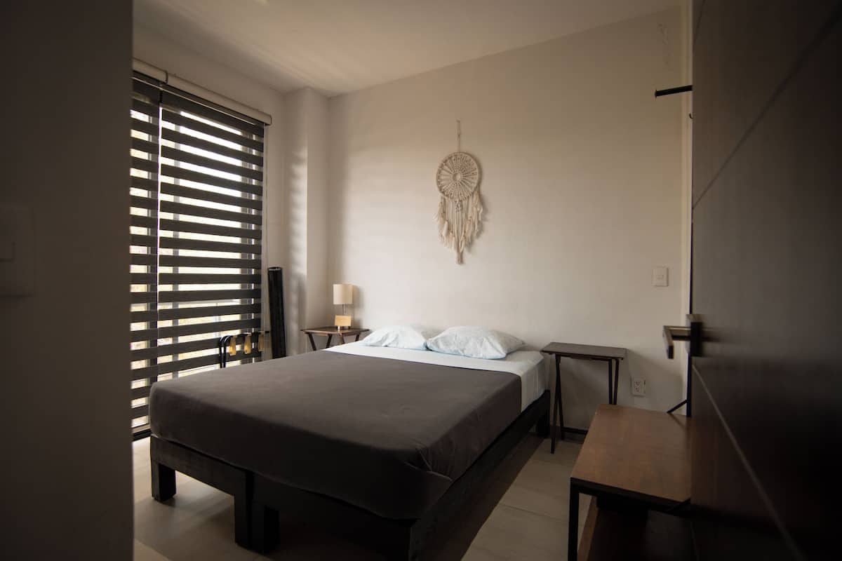 Un dormitorio con cama, mesitas de noche, una lámpara auxiliar y un atrapasueños colgado en la pared, además de una ventana con persianas.