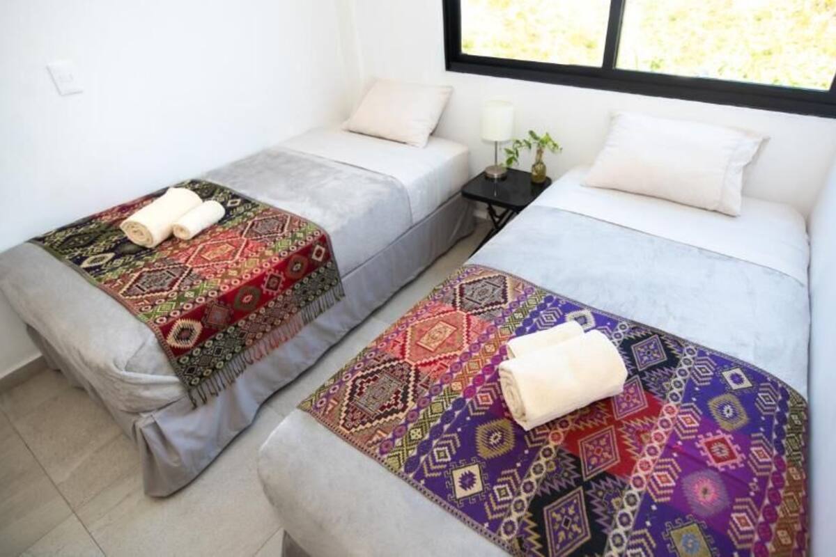 Dos camas individuales con cubrecamas estampados, una mesita de noche con lámpara y una ventana con vistas a zonas verdes.