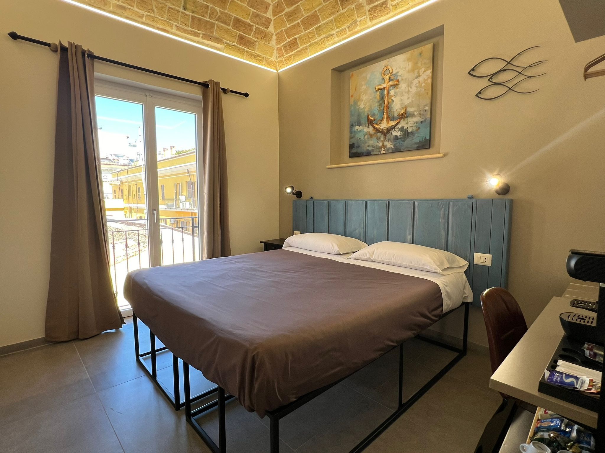 A hotel room with a bed, a desk with a chair, a painting, and a view of buildings outside.