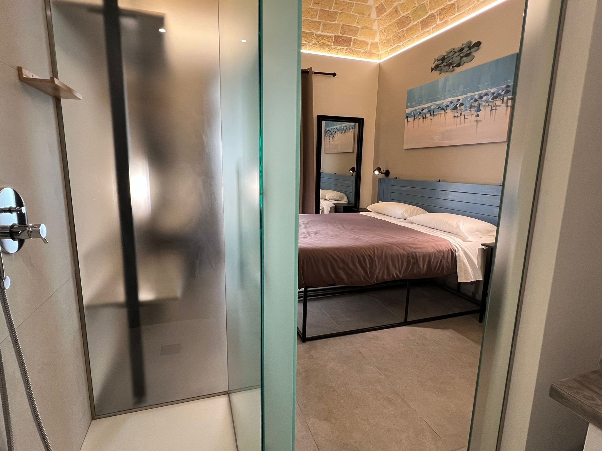 A modern bathroom with a glass shower enclosure, a visible toilet, and a bed with a blue headboard.