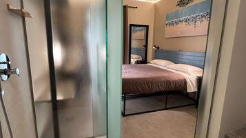 A modern bathroom with a glass shower enclosure, a visible toilet, and a bed with a blue headboard.