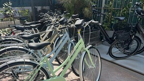 Filas de bicicletas estacionadas en un espacio al aire libre con piso de baldosas, rodeado de vegetación y una cerca.