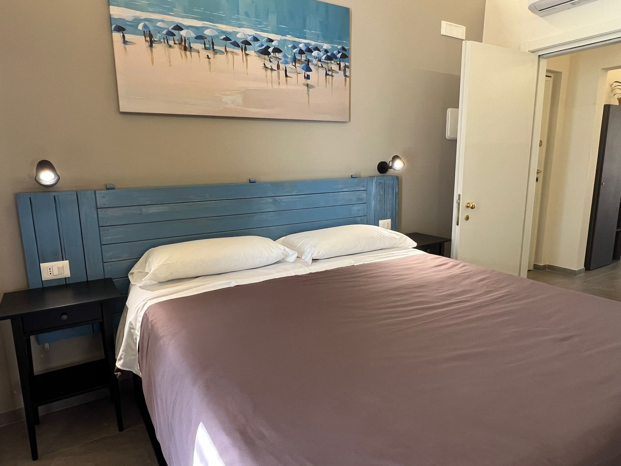 A bedroom with a blue headboard, a bed with white pillows, a nightstand, and a painting of a beach scene on the wall.