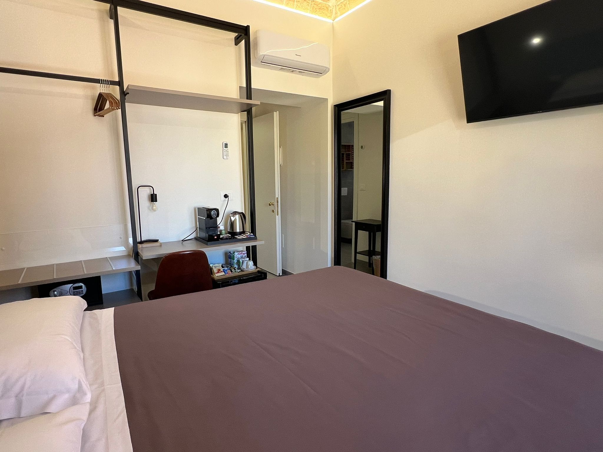 Ein Hotelzimmer mit einem Bett, einem Schreibtisch mit Kaffeemaschine, einem Sessel, einem an der Wand befestigten Fernseher und einer Tür zu einem Badezimmer.
