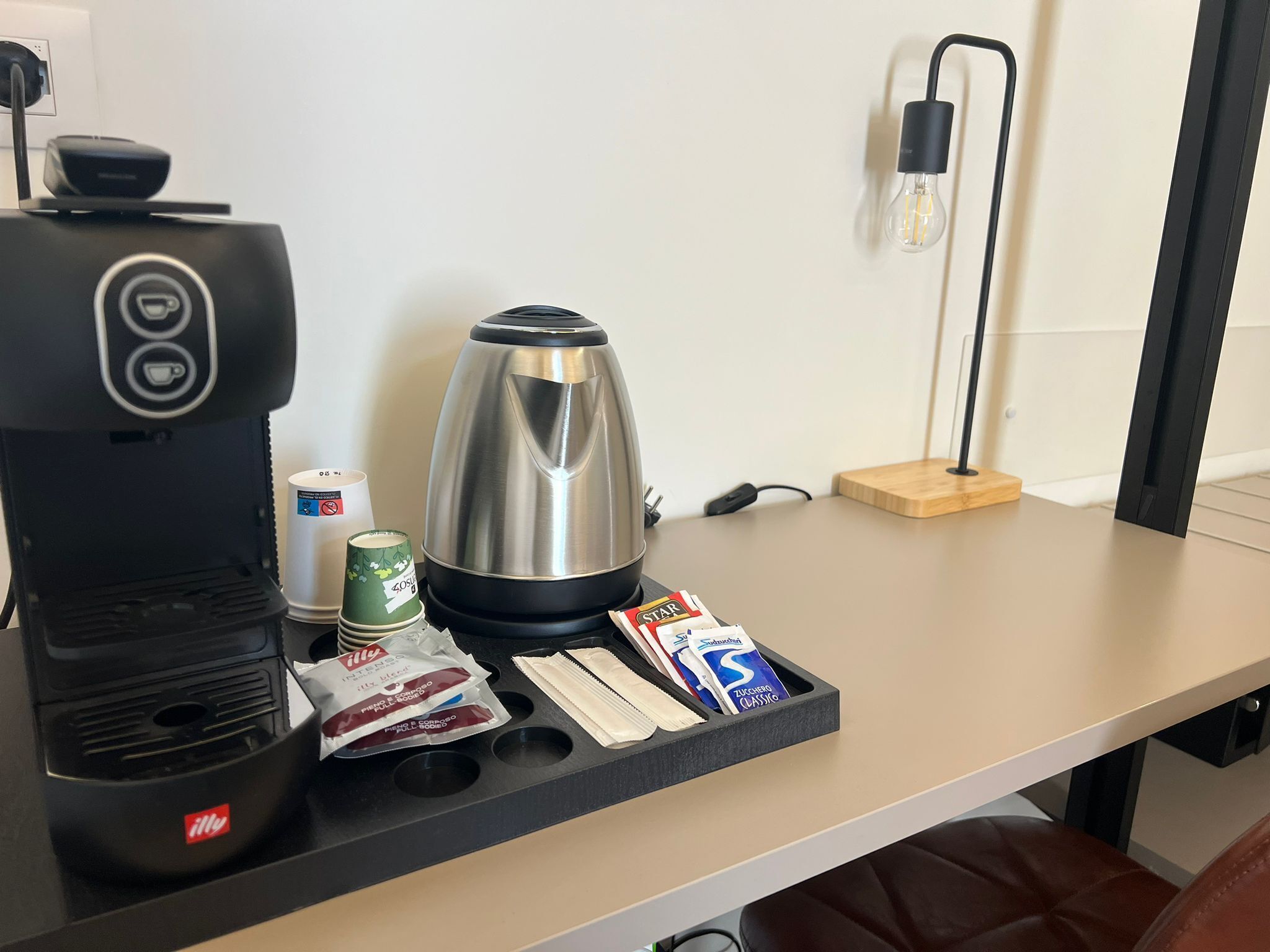 En kaffebryggare, en vattenkokare, en kopp kaffe, ett paket socker, ett paket grädde och ett paket kaffekapslar, allt placerat på ett bricka.