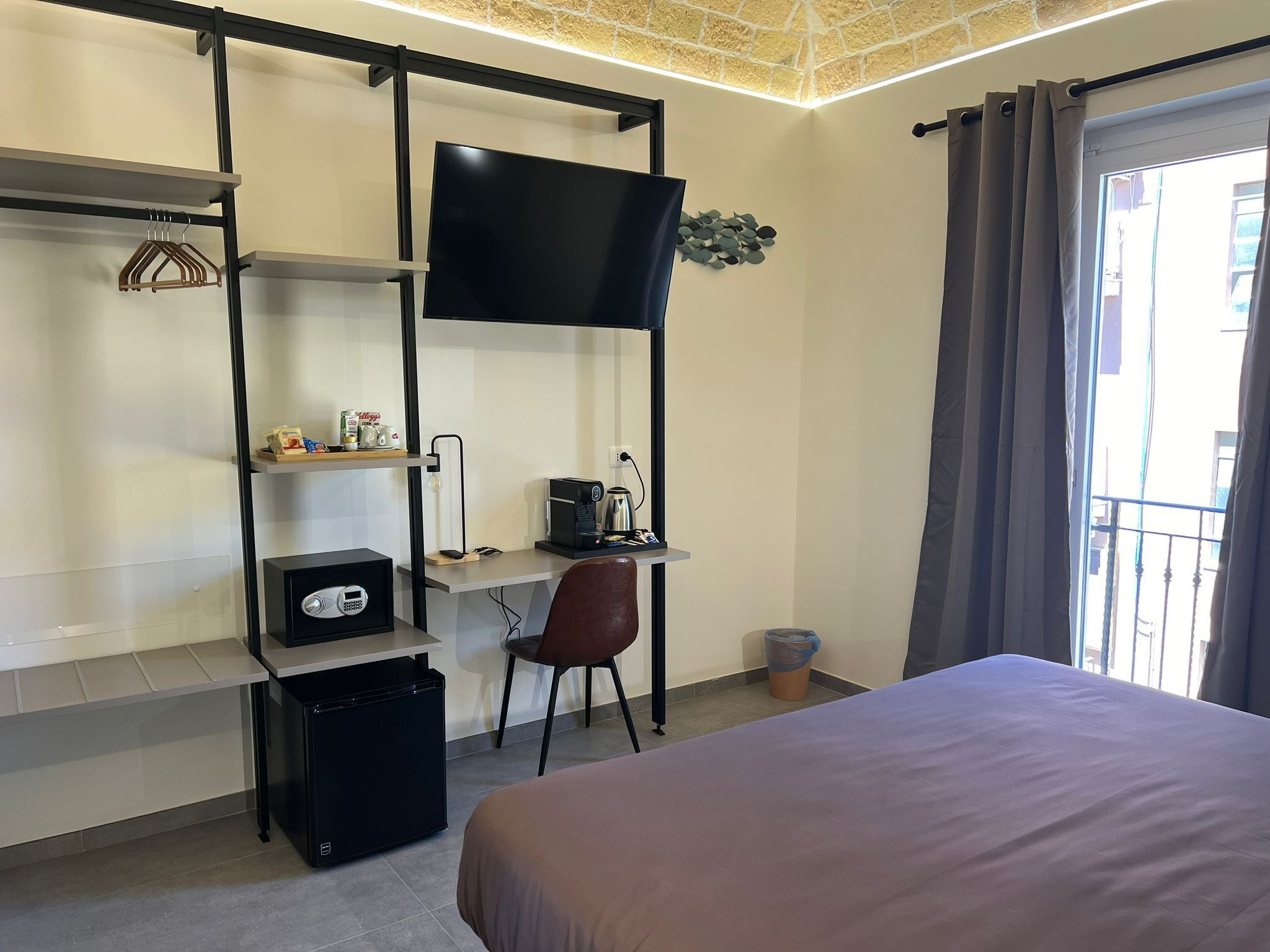 Une chambre d’hôtel de taille réduite, comprenant un lit, un bureau avec un ordinateur et une machine à café, une télévision et une petite poubelle.