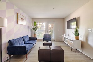 Un salon avec un canapé bleu, un pouf marron, un fauteuil bleu, un téléviseur fixé au mur et une fenêtre avec des stores.