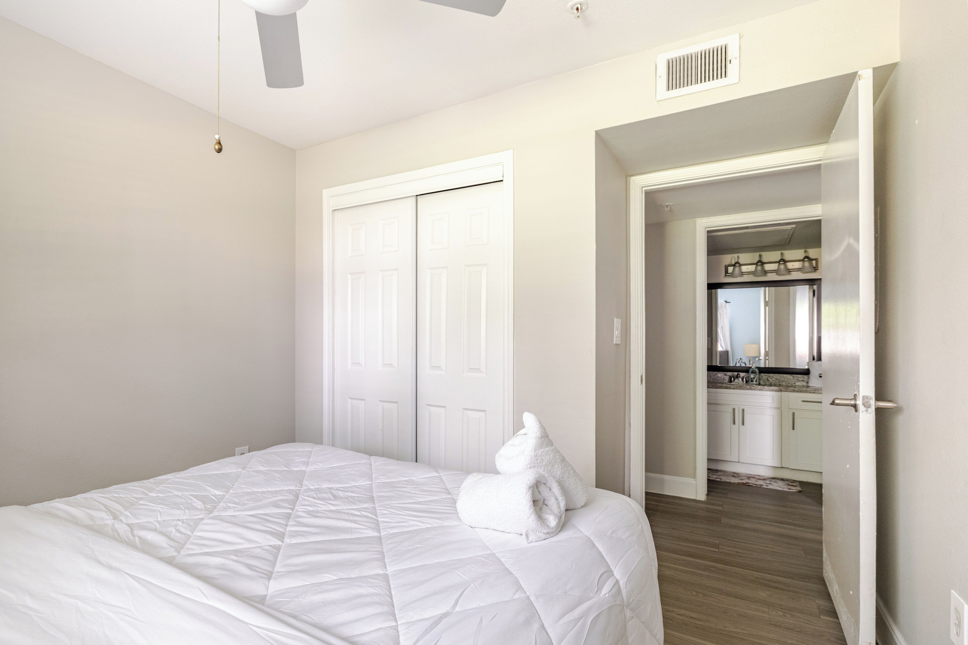 A bedroom with a bed, a white towel, a ceiling fan, and a door leading to a kitchen.