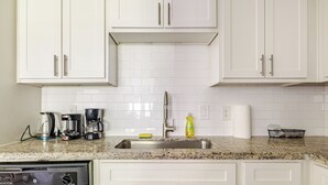 Una cocina con armarios blancos, encimera de granito, lavavajillas, cafetera, jarra de café, botella de detergente para lavar trastes y dispensador de toallas de papel.