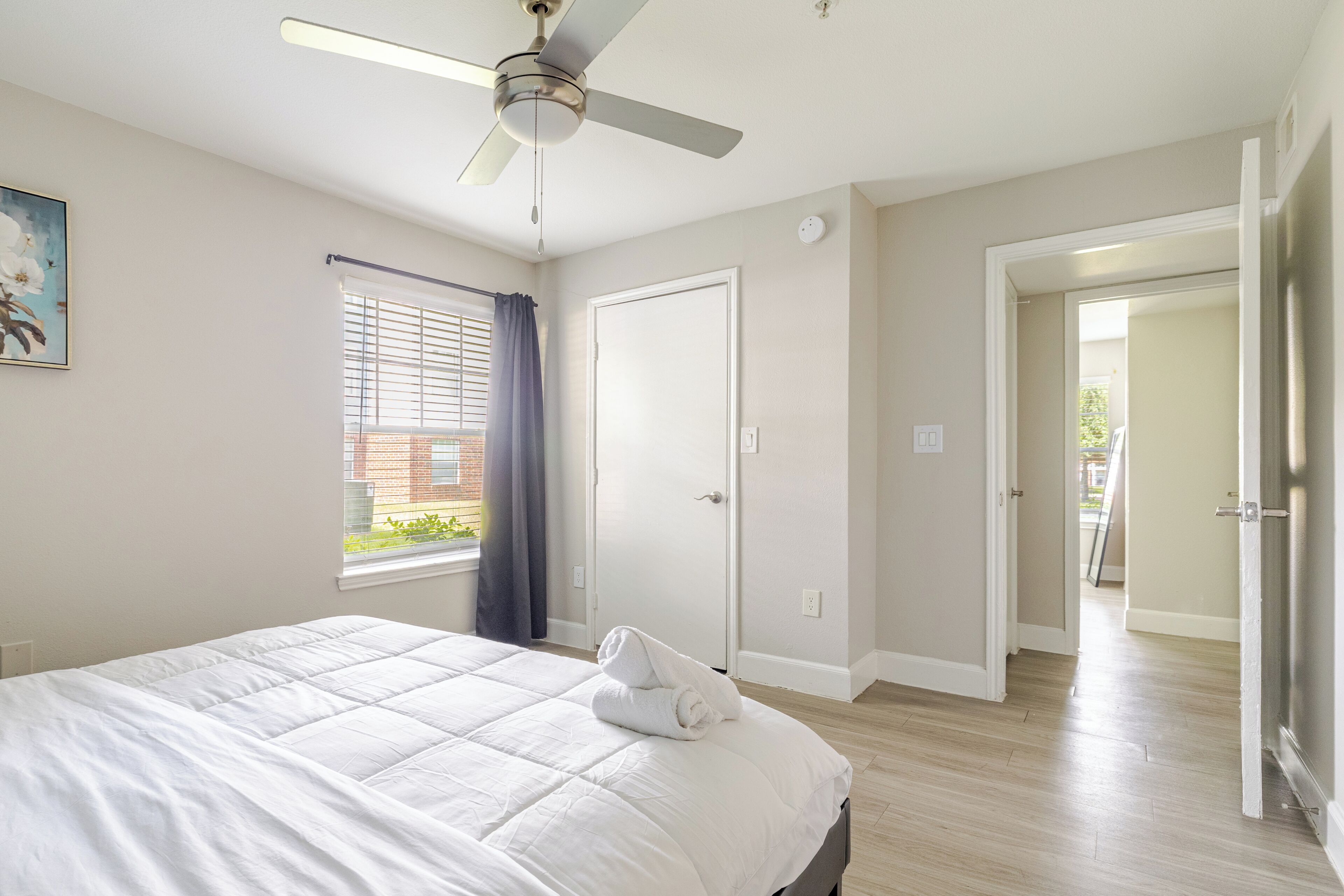A bedroom with a bed, ceiling fan, window with blinds, and a door leading to another room.