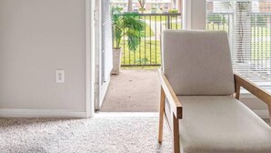 Una habitación con piso alfombrado, una silla de madera, una puerta que da a un área exterior con vista a un árbol y una valla.