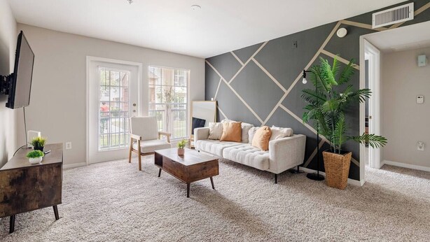 Una sala de estar con un sofá, una mesa de centro y un televisor. Hay ventanas con persianas, una planta y una pared con diseños geométricos.