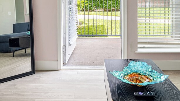 A room with a large mirror, a black coffee table with a colorful glass object, and a view of a brick building through a window with blinds.