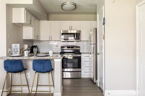 A modern kitchen with white cabinets, stainless steel appliances, and a granite countertop. There are two blue bar stools and a stainless steel refrigerator.