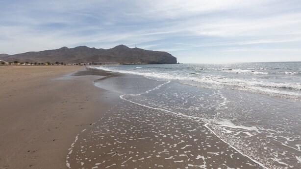 Una playa con una montaña al fondo, olas tranquilas y cielo despejado.