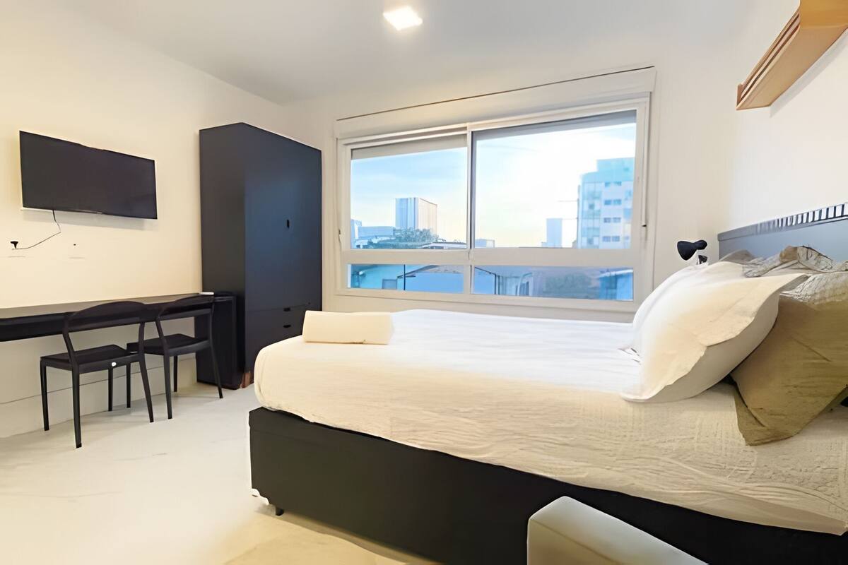Une chambre avec un lit, un bureau, une chaise, un téléviseur fixé au mur et une grande fenêtre donnant sur la ville.