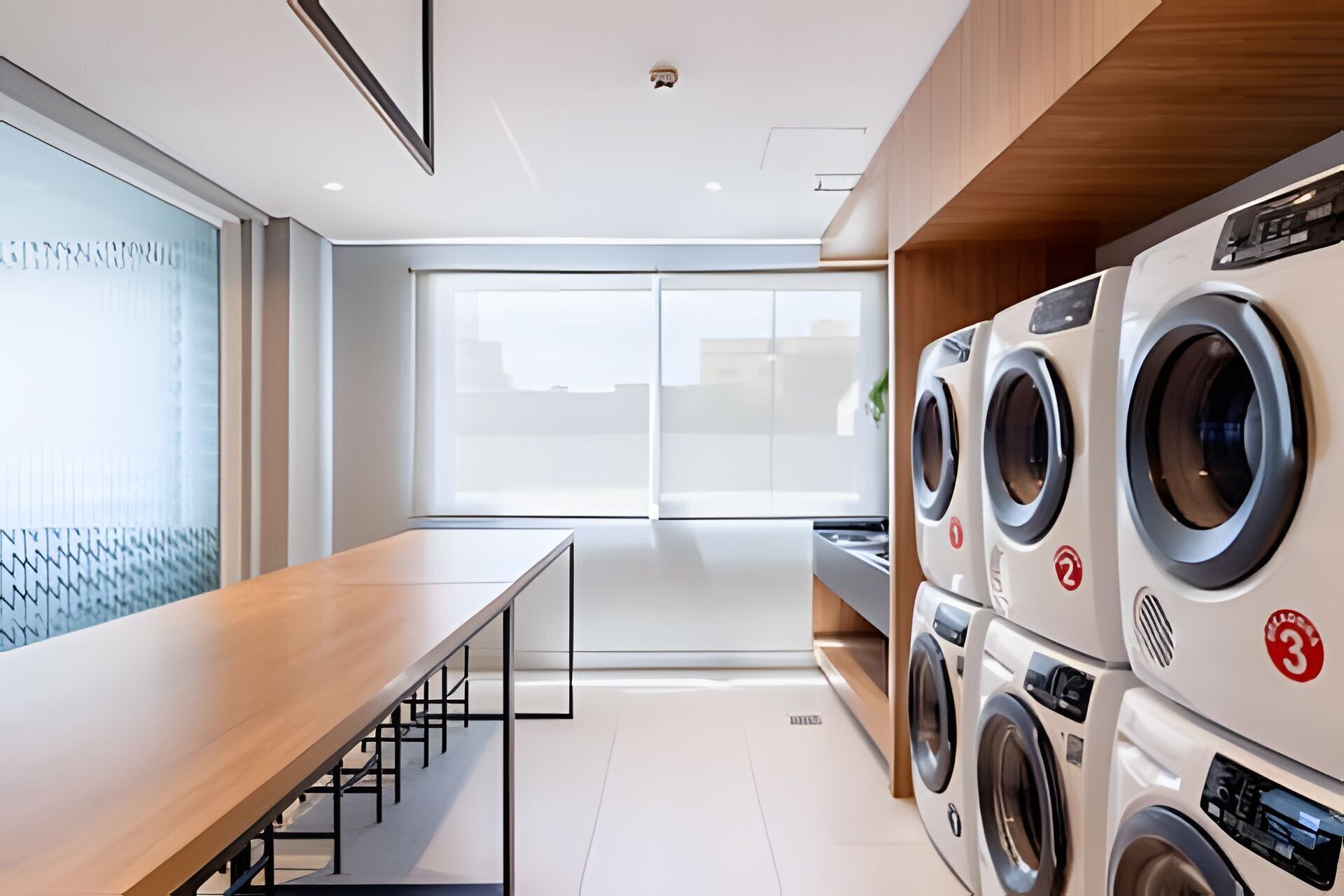 Un lavadero moderno con un banco de madera largo, ventanas amplias y lavadoras de carga frontal apiladas.