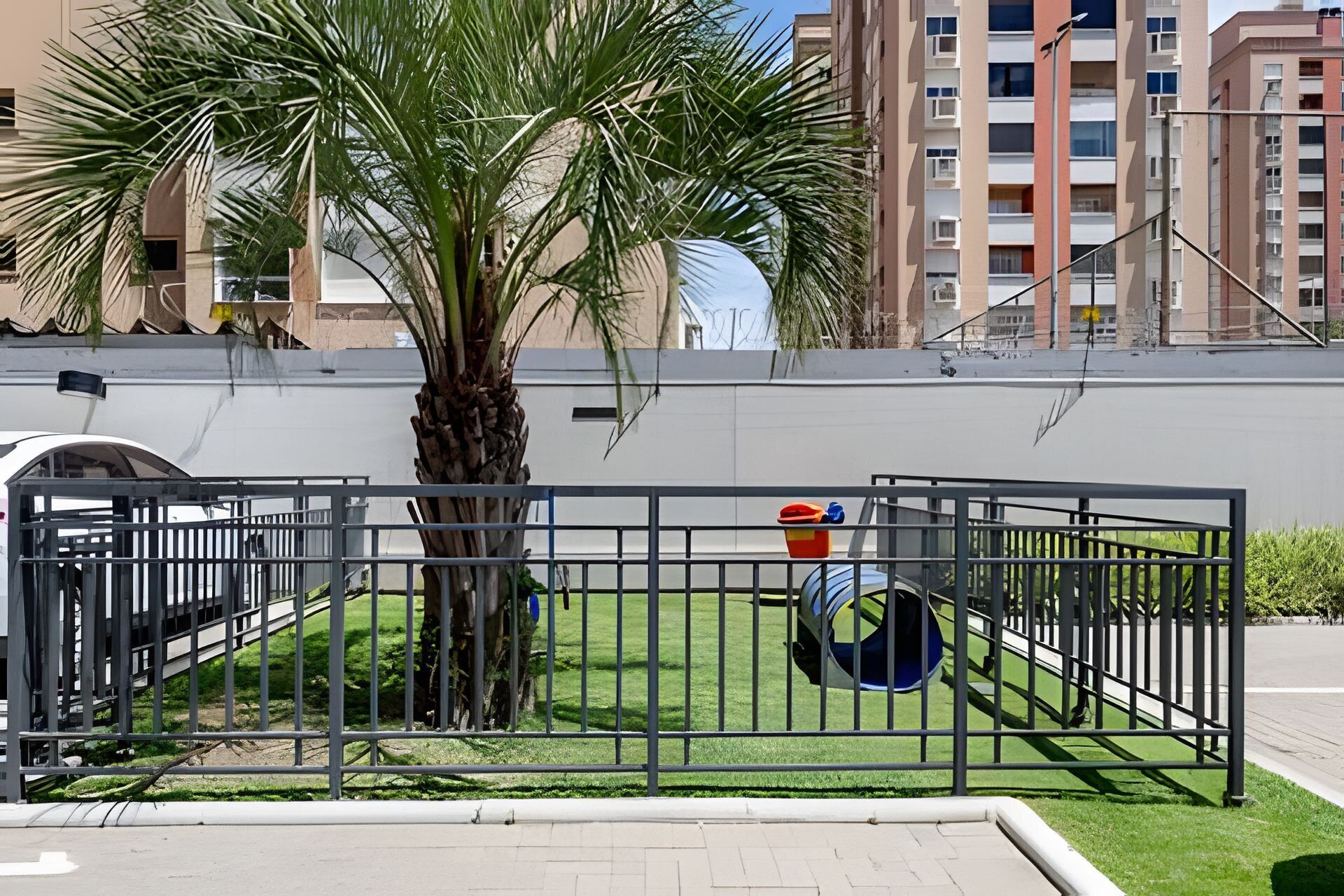 A fenced outdoor area with a palm tree, a playground with a slide, and a building in the background.