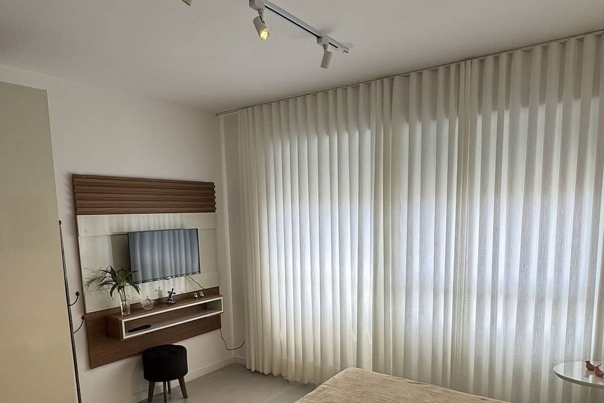 Une chambre d’hôtel avec un grand lit, un téléviseur fixé sur un panneau en bois, un bureau avec une chaise et une fenêtre équipée de stores verticaux.