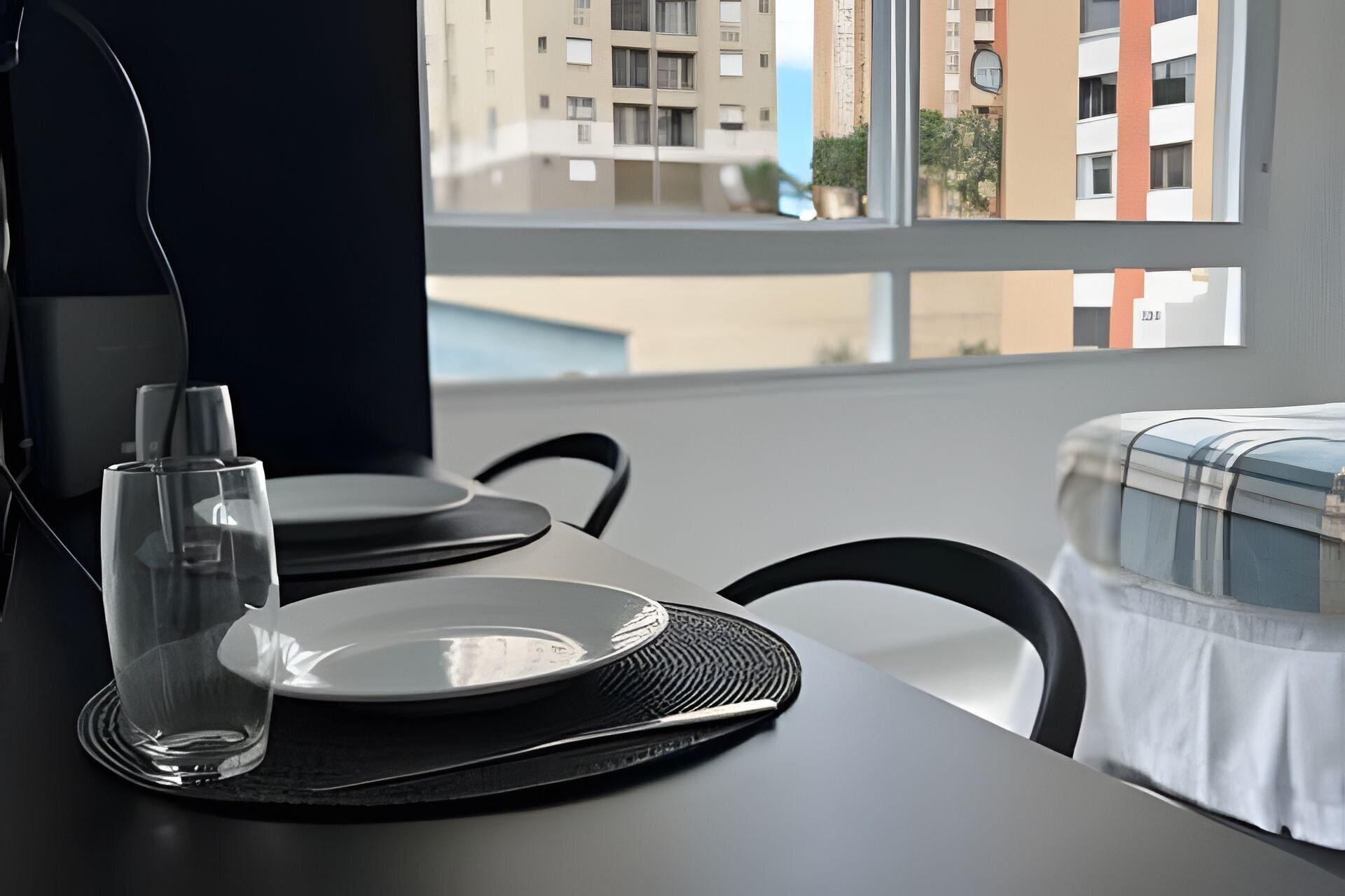 A dining table with a glass, plate, and black placemat. A window reveals a cityscape with buildings.